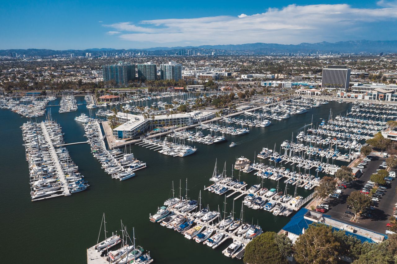 Wide aerial view of Marina del Rey harbor at golden hour, boats in slips, Ballona Creek and the Pacific in the background, descriptive alt text: "Aerial view of Marina del Rey harbor filled with sailboats and yachts at sunset"